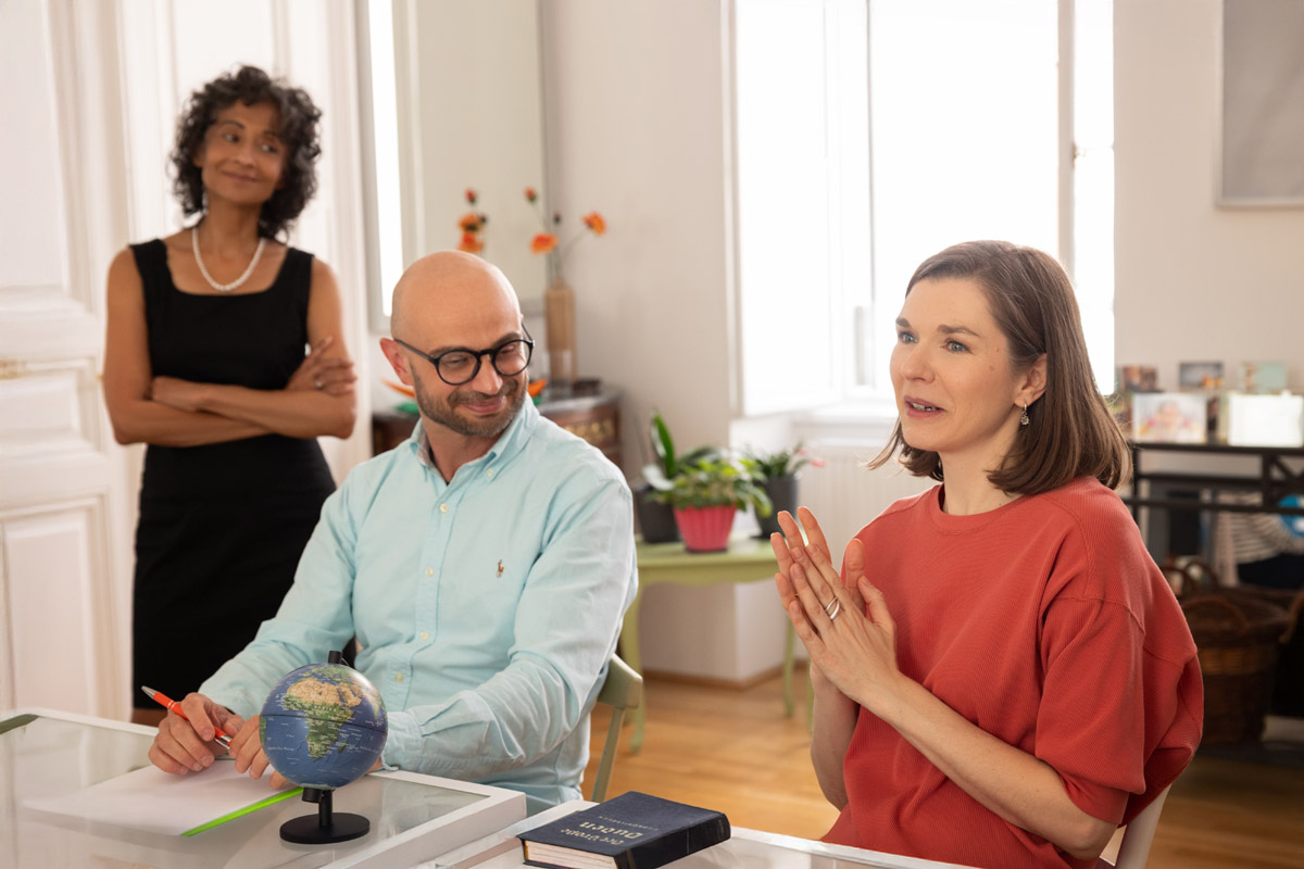 Eine Workshopteilnehmerin spricht neben einem lächelnden Teilnehmer, mit Michèle Cooke im Hintergrund stehend.